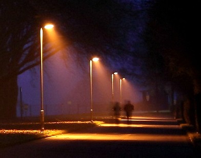 Two people walking down a darkened street lit only by a few lamp posts