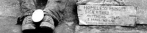 The image of a homeless man's feet, an empty cup between, and a sign that says, I have no one.