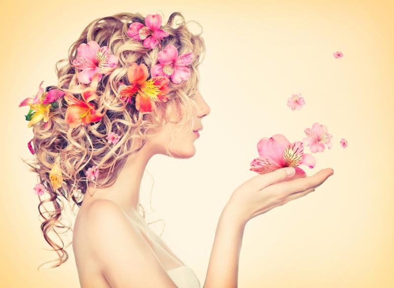 Beauty girl takes beautiful flowers in her hands. Blowing flower. Hairstyle with flowers.  Fantasy girl portrait in pastel colors. Summer fairy portrait. Long permed hair.