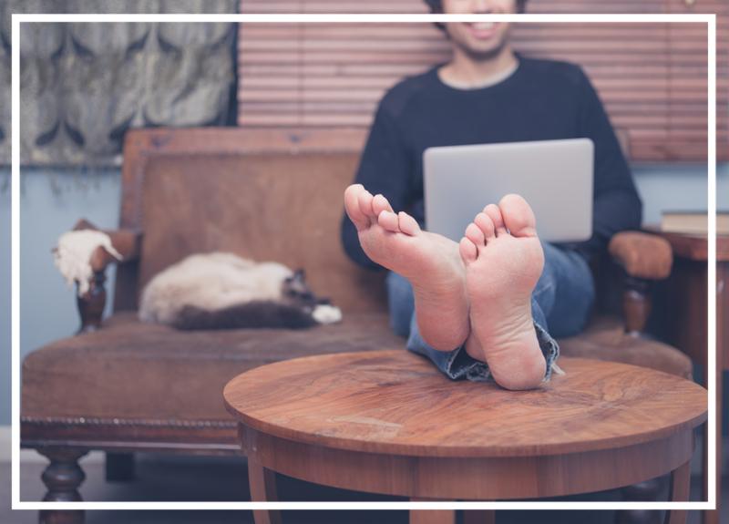 A happy barefoot man is resting his feet on a coffee table at home while working on his laptop there is a cat sleeping next to him