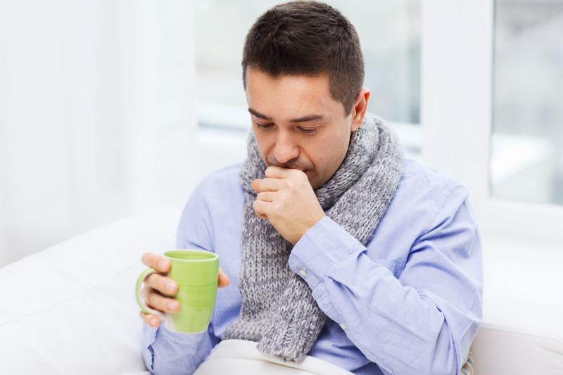 healthcare, people and medicine concept - ill man with flu coughing and drinking hot tea from cup at home