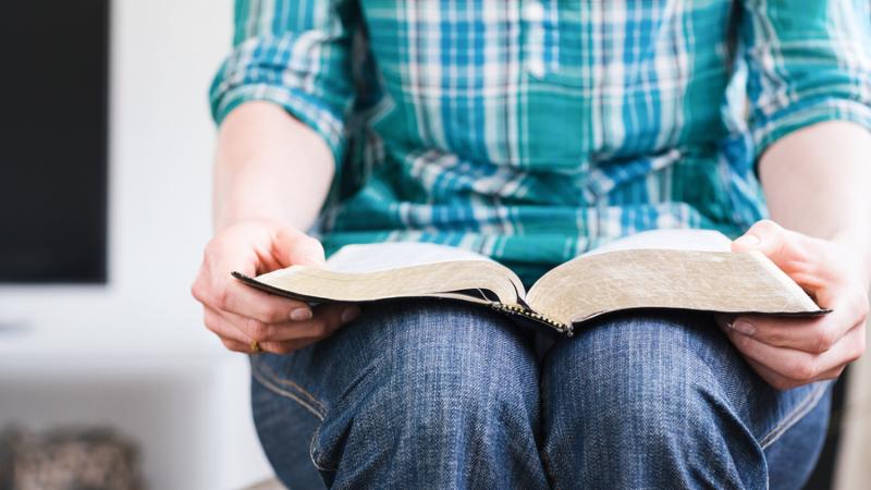 A young woman at home holds an open Bible in her lap. Closeup image with shallow DOF focus on the tip of the book.
