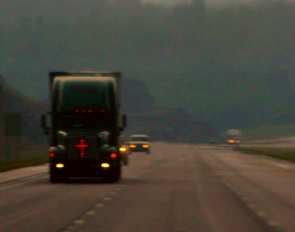 A semi truck, driving in the rain with a lighted cross on its grill