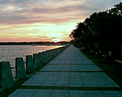 Empty walkway beside a calm river leading to the rising sun