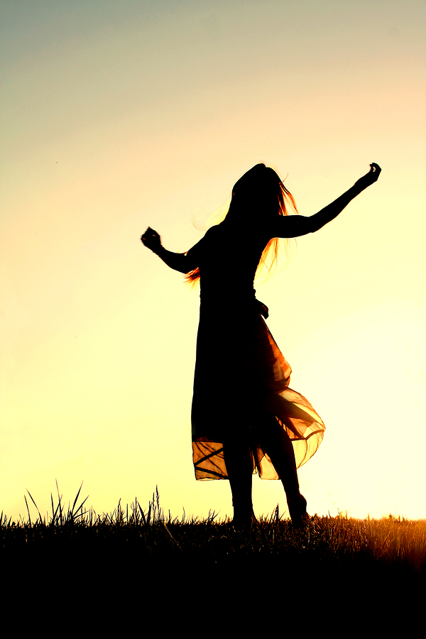 A woman wearing a long skirt with long blonde hair is dancing and praising God while silhouetted against the evening sky